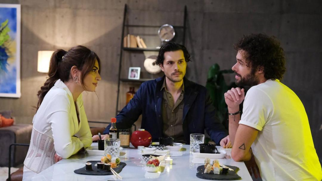 A imagem mostra um homem sentado à frente de uma mulher, numa mesa de jantar e outro homem olhando os dois