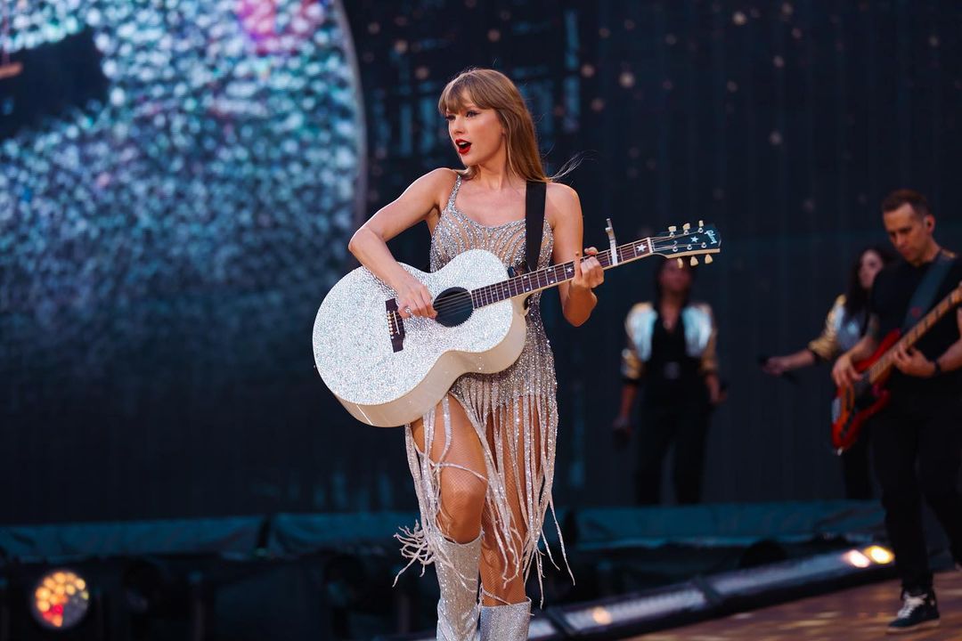 A imagem mostra Taylor Swift, uma mulher branca, de cabelos loiros, lisos e longos, cantando durante um de seus shows da turnê 