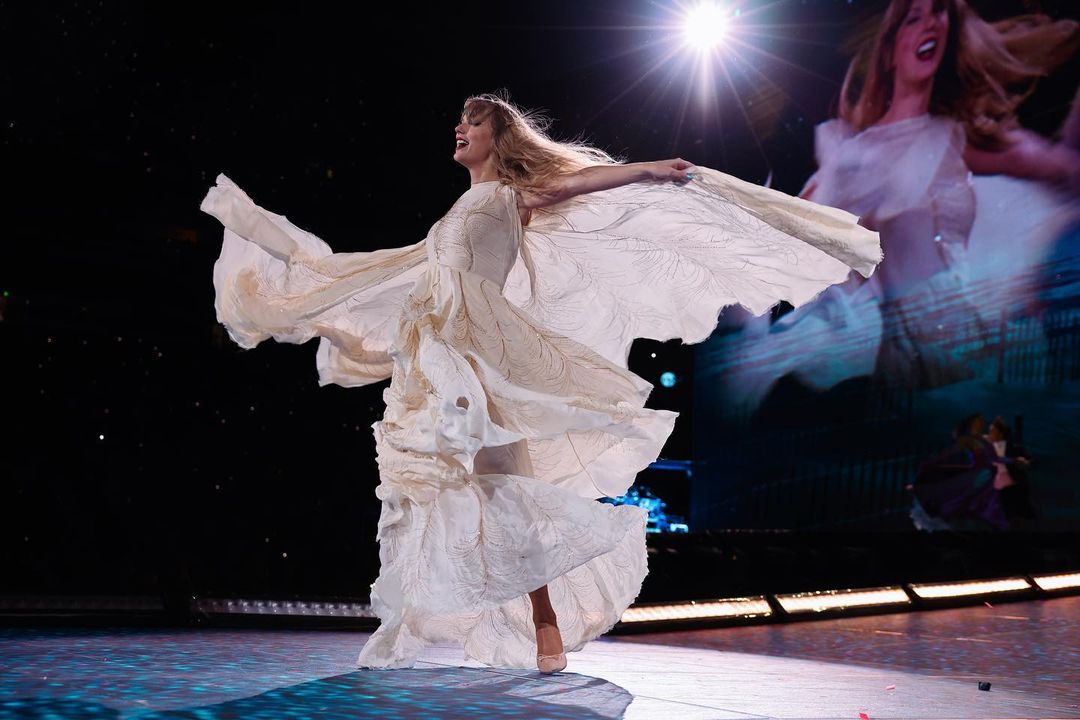 A imagem mostra Taylor Swift, uma mulher branca, de cabelos loiros, lisos e longos, cantando durante um de seus shows da turnê 