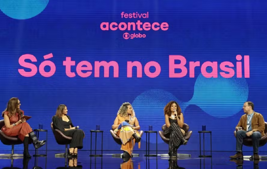 Imagem mostra um grupo de cinco pessoas sentadas e conversando no Festival Acontece. Atrás, em um fundo azul, há os dizeres do festival junto da seguinte frase 