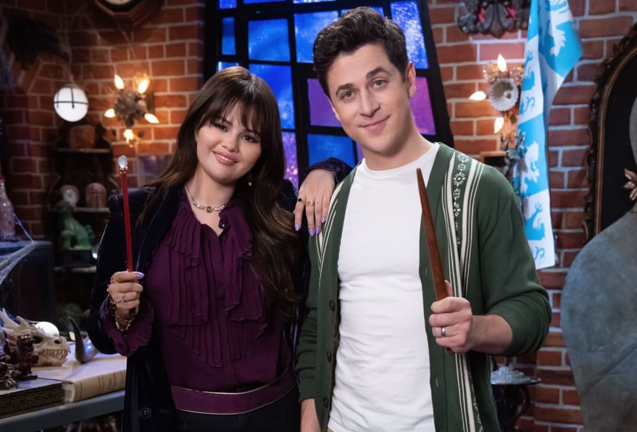 Imagem mostra Selena Gomez segurando uma varinha de magia junto a David Henrie. Eles posam para foto atrás do cenário da série Os Feiticeiros de Waverly Place