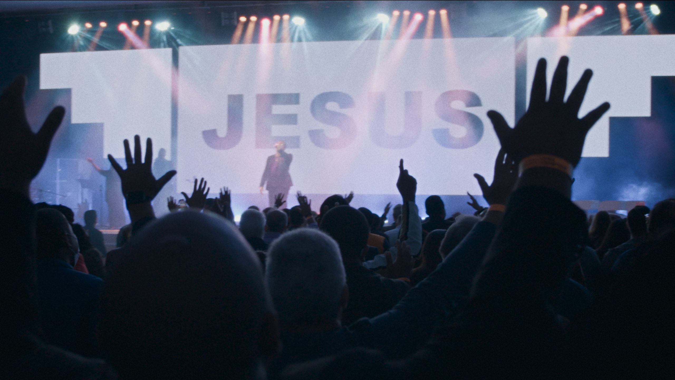 Telão com o texto "Jesus" e pessoas com as mãos para cima.