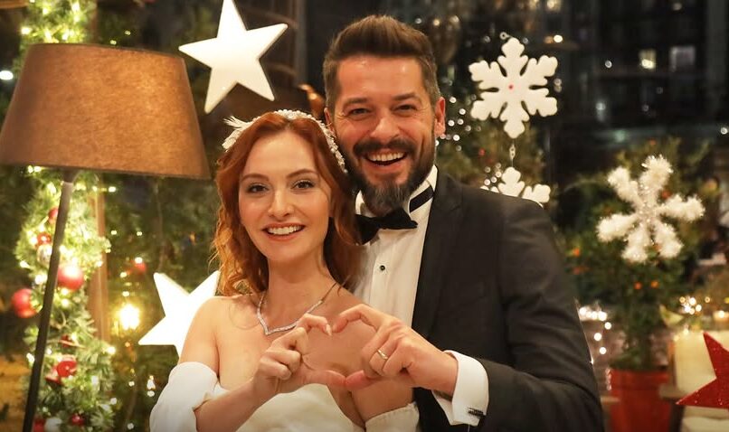 Foto dos personagens Piril e Engin de “Sen Çal Kapımı” fazendo um coração, com decorações natalinas ao fundo.