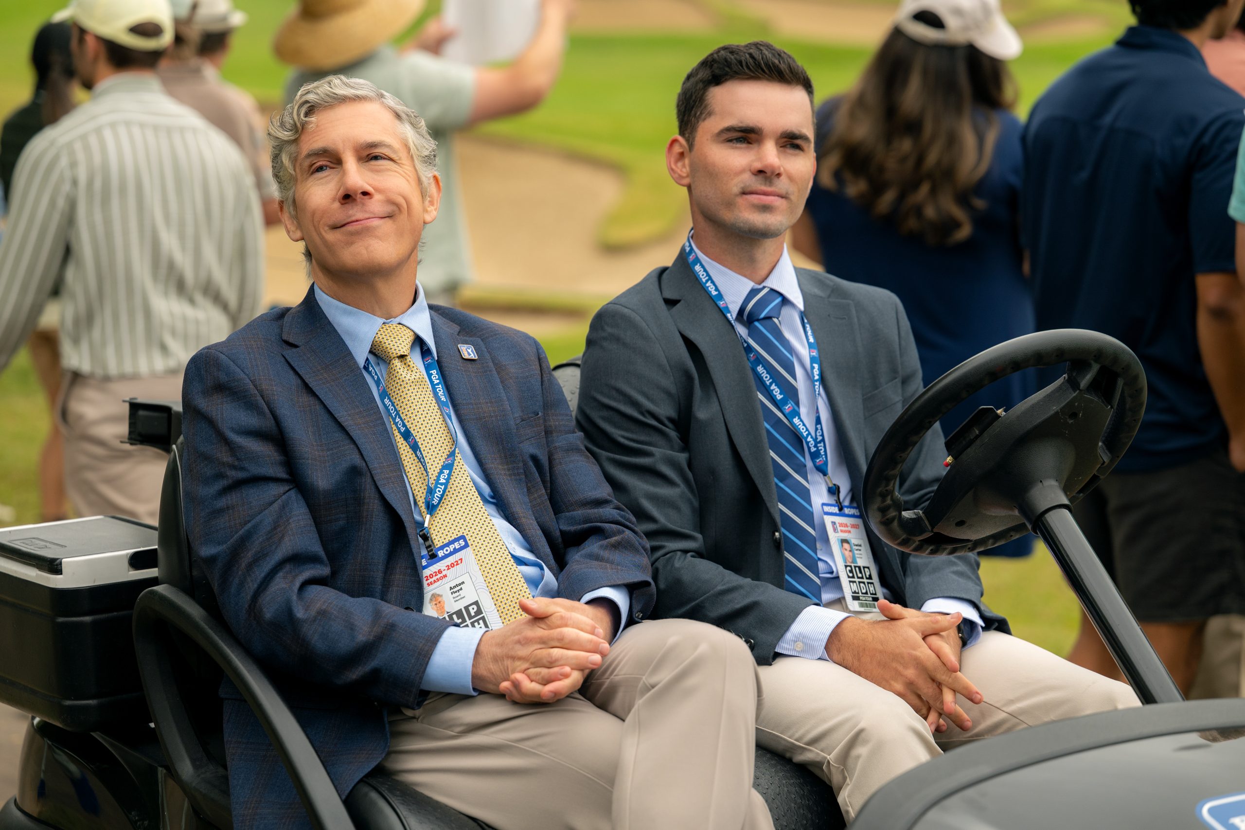 "Two men in suits sit in a golf cart at a golf course event, wearing event badges, with several people standing in the background on a grassy area near sand traps. The setting appears formal and outdoors during daytime."