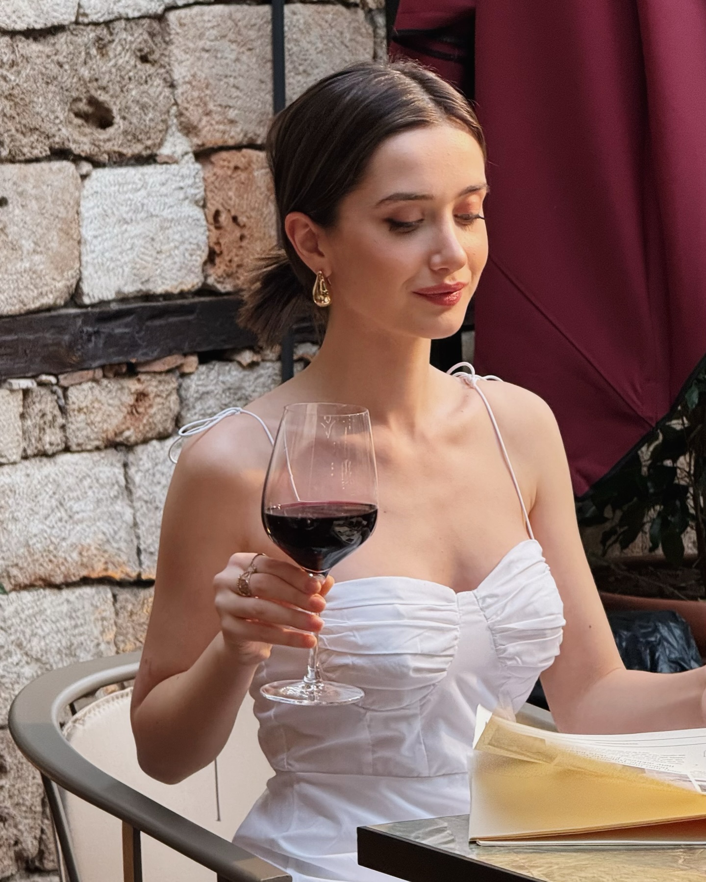 İlsu Demirci sentada enquanto lê algumas folhas. Ela está segurando também uma taça de vinho e usando uma blusa/vestido branco.