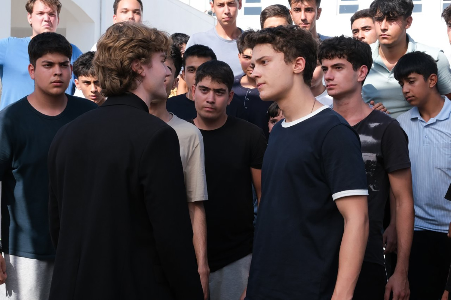 Cena de Daha 17 em que dois garotos vestidos de preto se encaram, enquanto alguns jovens aparecem ao fundo.