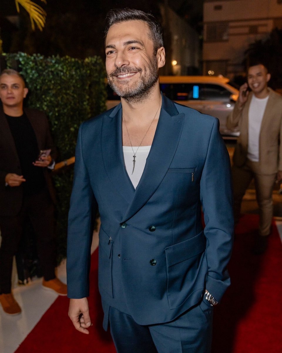 Barış Kılıç, usando um terno azul, sorri enquanto caminha sobre um tapete vermelho durante um evento noturno.