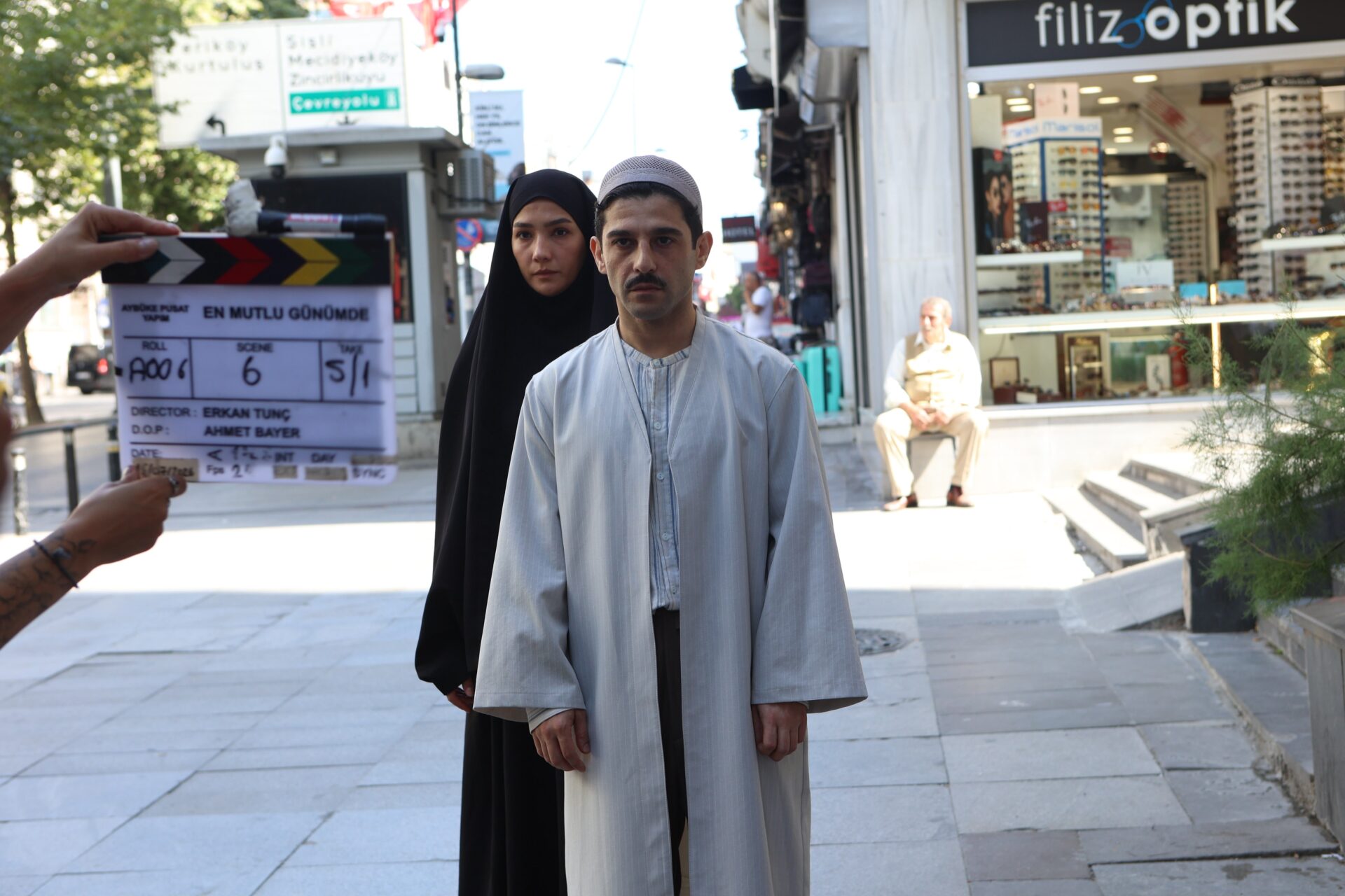 Halil İbrahim Babür vestido com roupas tradicionais e Aybüke Pusat usando um hijab aparecem durante as gravações de En Mutlu Günümde em uma rua comercial.