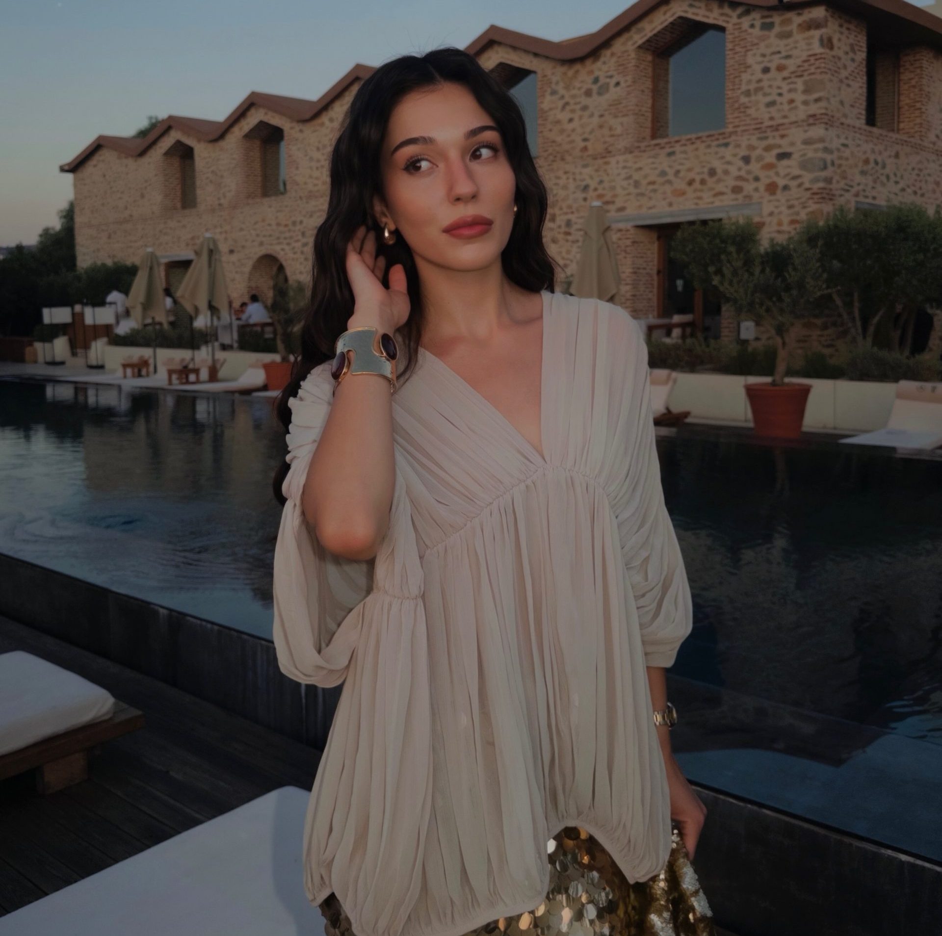 Meltem Akçöl usando um vestido branco e posando ao lado de uma piscina ao entardecer, em frente a um conjunto de construções de pedra.