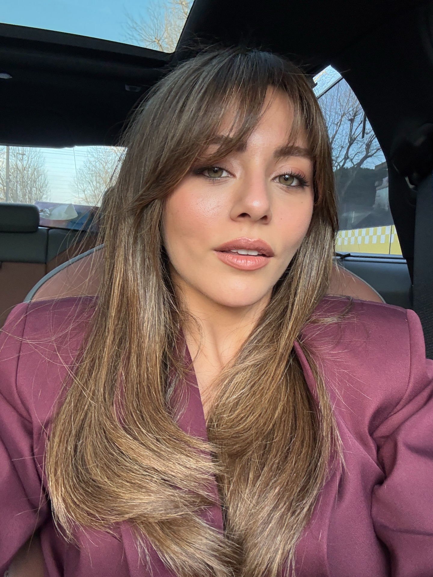 Sıla Türkoğlu de cabelos castanhos posa para uma selfie dentro de um carro, usando um blazer vinho.
