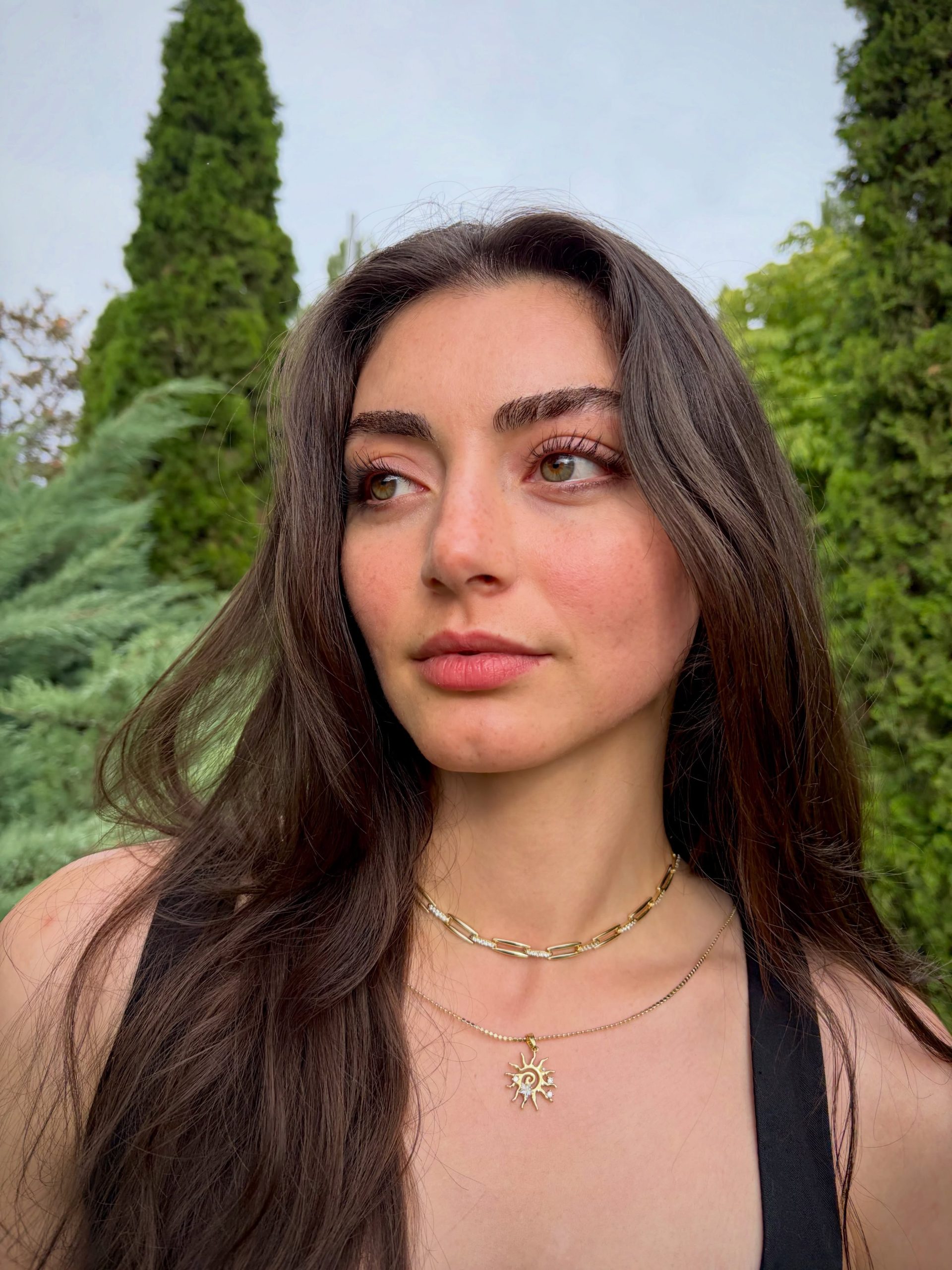 Özge Törer com cabelos longos e escuros posando ao ar livre, usando colares dourados, com árvores ao fundo.