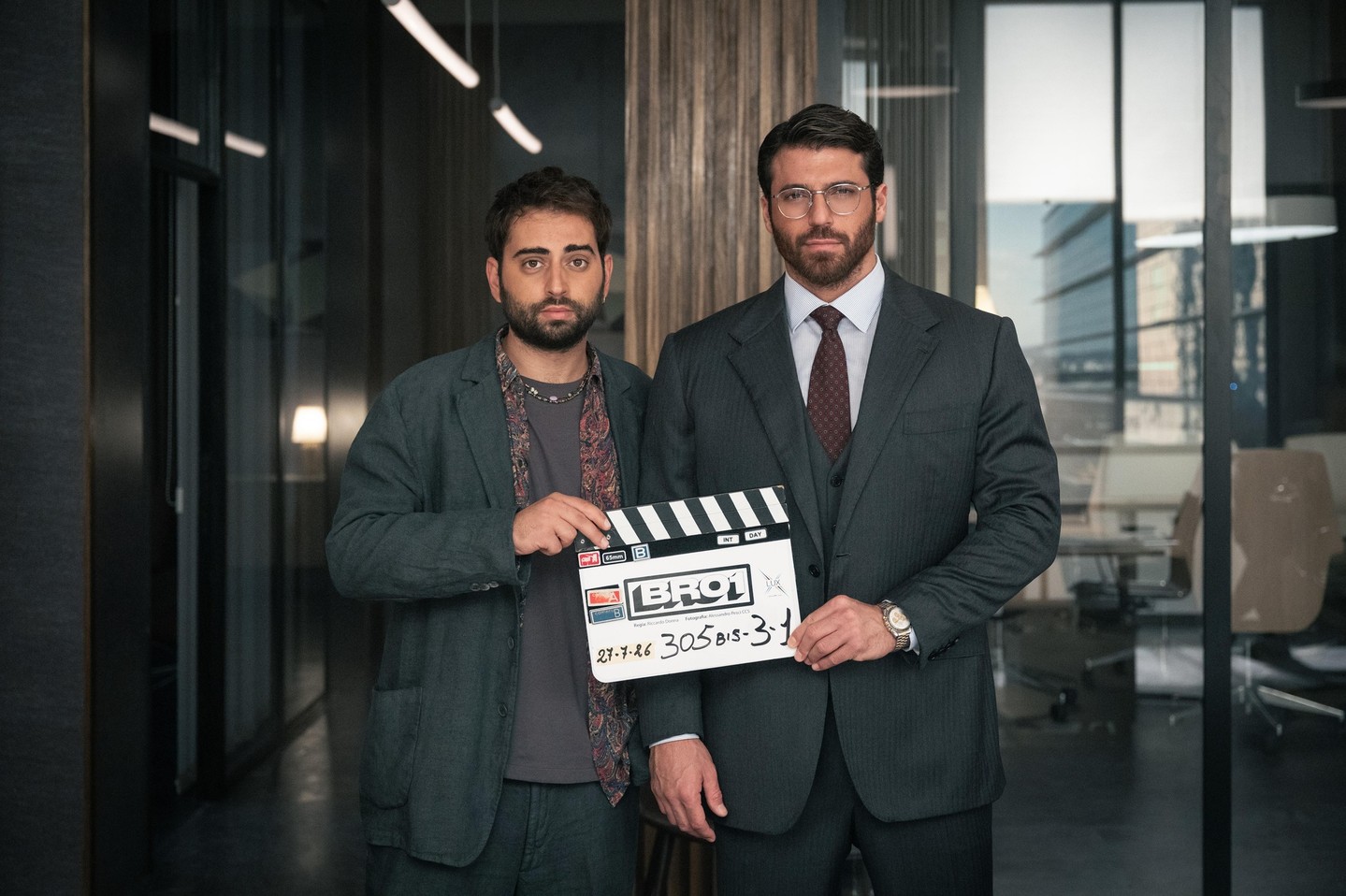 Giovanni Nasta e Can Yaman posando lado a lado em um escritório, segurando a claquete de gravação da série Bro.
