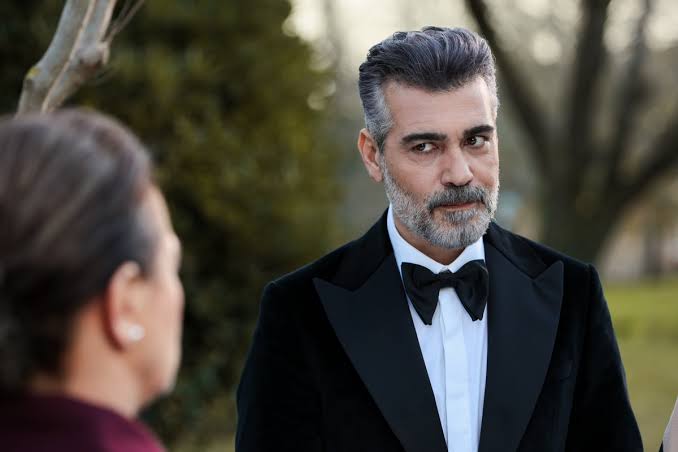 Personagem de Caner Cindoruk usando um terno preto, enquanto conversa com outra personagem.