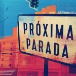 Uma placa com o nome do podcast "Próxima Parada" está em destaque em primeiro plano. Ao fundo há algumas construções e um semaforo.