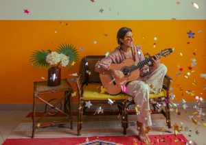 Imagem de Pe Lu sentado em um sofá com um violão e confetes de carnaval caindo