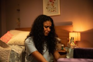 Uma mulher de cabelos cacheados está sentada no chão de um quarto, apoiada na cama, enquanto chora encarando uma garrafa quase vazia.