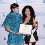 Sofia Carson está ao lado de Valentina García, a jovem beneficiada com a bolsa. As duas seguram juntas um documento oficial sobre a bolsa e estão posicionadas na frente de um painel escrito “Latin GRAMMY Cultural Foundation”. Valentina está sorrindo enquanto olha para câmera e Sofia está sorrindo enquanto olha para o lado.