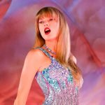 A imagem mostra Taylor Swift, uma mulher branca, de cabelos loiros, lisos e longos, cantando durante um de seus shows da turnê "The Eras".