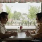 Barbara Ferreira e John Leguizamo de frente um para o outro em uma mesa.