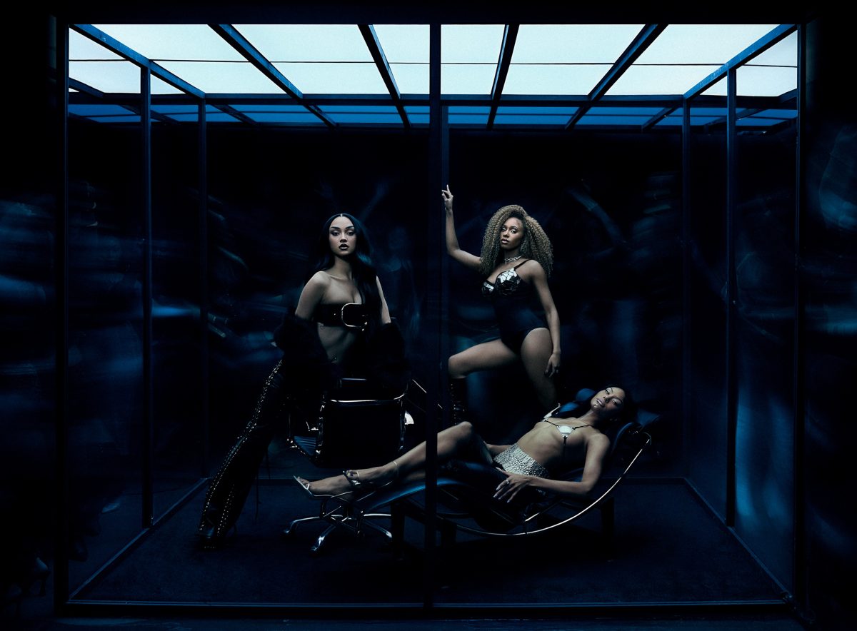 Foto conceitual do trio feminino britânico FLO em um cenário escuro e futurista. As três integrantes aparecem dentro de uma estrutura de vidro iluminada por um teto quadriculado em tons de azul frio, lembrando uma sala ou cubo transparente. À esquerda, uma integrante de cabelos longos e pretos posa em pé ao lado de uma cadeira moderna. Ela usa um look preto brilhante com recortes e detalhes metálicos, além de botas altas. No centro, outra integrante, com cabelos volumosos e cacheados, está em posição elevada, apoiando uma das mãos em uma coluna do cubo. Ela veste um body preto com detalhes prateados refletivos e botas de cano alto. À direita, a terceira integrante aparece deitada de forma elegante em uma chaise longue preta, usando um conjunto brilhante prateado e preto. Sua pose transmite relaxamento e confiança. A iluminação azulada cria um clima sofisticado, misterioso e cinematográfico. O fundo é quase totalmente escuro, com sombras e borrões ao redor do vidro, dando sensação de movimento e profundidade. A composição transmite luxo, poder e estética futurista.
