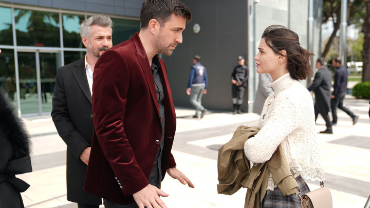 Kadir de terno vinho conversa com Meryem vestida de branco em frente a um edifício.
