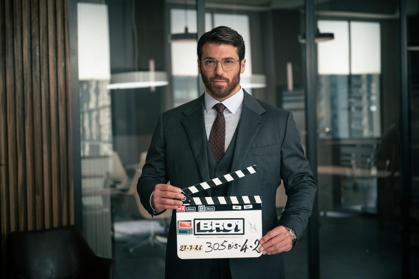 Can Yaman utilizando um terno em um escritório moderno, segurando uma claquete marcando o início da gravação da série Bro.