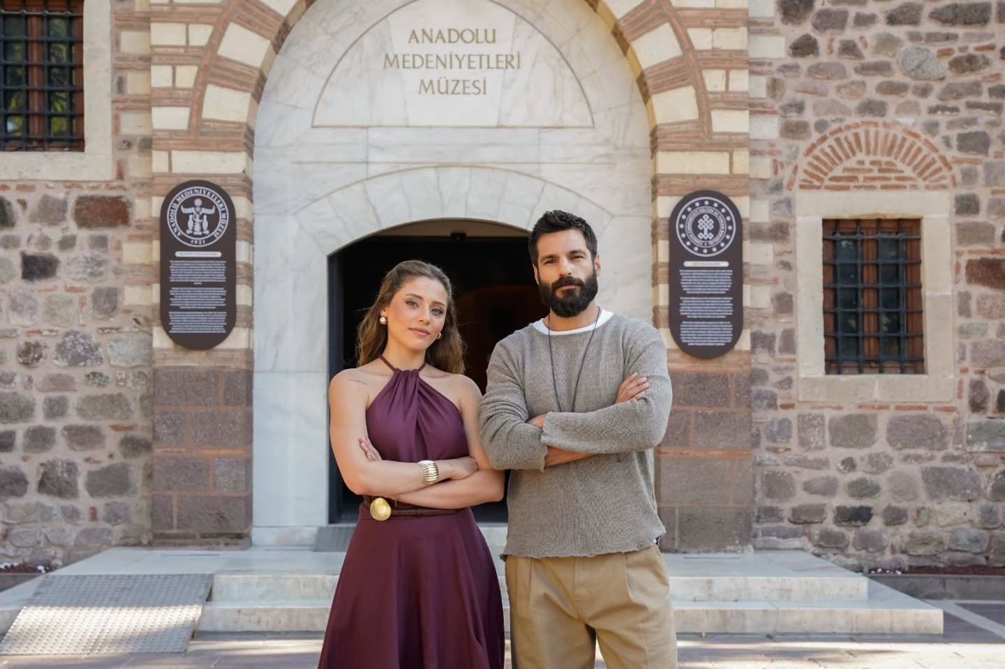 Imagem de Sıla Türkoğlu e Serkan Çayoğlu na frente do Museu das Civilizações da Anatólia. Ela usa um vestido vinho e ele está usando uma blusa cinza e uma calça cumprida bege.