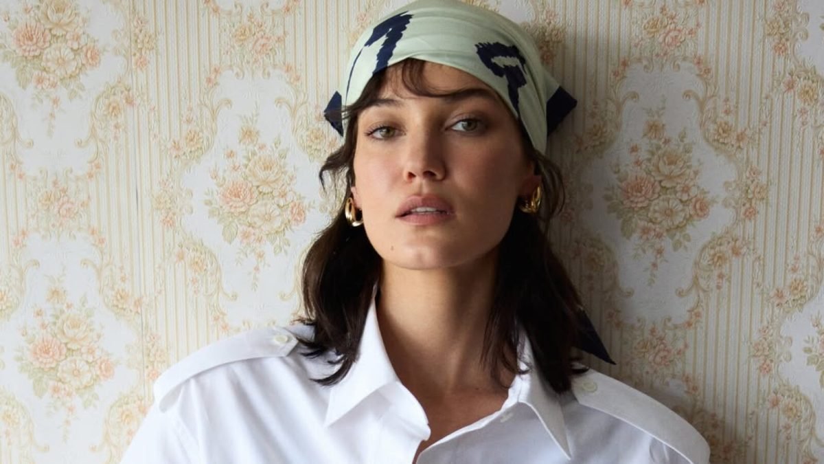 Pınar Deniz posa para um retrato usando uma camisa branca e um lenço estampado na cabeça, em frente a uma parede com papel de parede floral vintage.