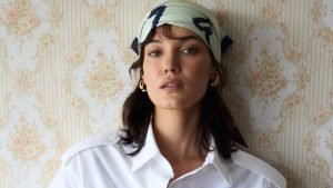 Pınar Deniz posa para um retrato usando uma camisa branca e um lenço estampado na cabeça, em frente a uma parede com papel de parede floral vintage.