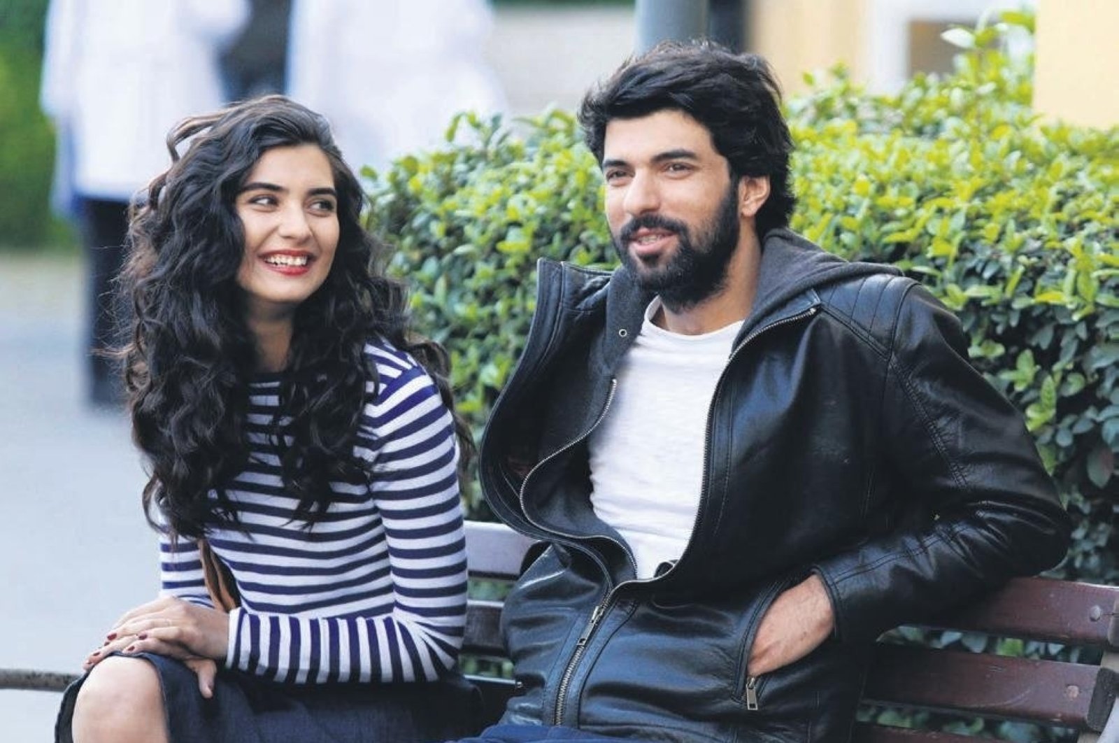 Personagens de Engin Akyürek e Tuba Büyüküstün aparecem sorrindo, sentados em um banco ao ar livre.