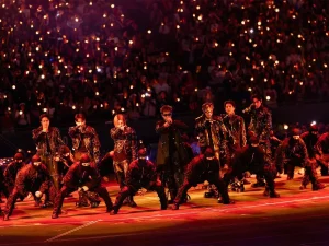 Vamos ser sinceros: por que sempre que os fãs latino-americanos reclamam de alguma coisa relacionada aos shows aparece alguém para dizer que eles deveriam agradecer pela oportunidade de ver o BTS? Parece que, para uma parte do fandom, morar na América Latina (América do Sul) significa ter que aceitar qualquer condição sem reclamar, como se receber uma turnê já fosse motivo suficiente para ficar quieto. E não, não é bem assim que funciona. É claro que organizar shows em diferentes países não é tão simples quanto parece. Existem diferenças de estrutura, logística, custos e várias outras questões que influenciam a produção de uma turnê. Ninguém está dizendo que todos os países precisam receber exatamente o mesmo palco ou que tudo funciona da mesma maneira em qualquer lugar. Mas reconhecer essas dificuldades é bem diferente de usar isso como argumento para invalidar toda reclamação dos fãs latinos. Afinal, por que alguém que compra um ingresso na América Latina deveria ter menos liberdade para questionar a experiência que está pagando para viver? Um fã brasileiro, mexicano, chileno ou argentino não precisa provar que ama o BTS tanto quanto alguém dos Estados Unidos, da Europa ou da Coreia do Sul. Também não precisa mostrar quantos streams fez, quantos álbuns comprou ou quantas metas ajudou a bater para ter o direito de dizer que alguma coisa poderia ser melhor. Desde quando ser fã significa aceitar tudo sem reclamar? Essa é uma das coisas mais cansativas nessa discussão. Às vezes, parece que o ARMY latino precisa apresentar uma lista de tudo o que já fez pelo grupo antes de poder falar sobre qualquer problema. E, quando finalmente reclama, ainda corre o risco de ser chamado de ingrato, como se apontar algo que incomoda fosse o mesmo que não valorizar a presença do BTS na região. Mas dá para estar feliz com a possibilidade de ver os meninos e, ao mesmo tempo, querer uma organização melhor. Dá para esperar anos por um show, juntar dinheiro para comprar o ingresso e ainda questionar decisões que afetam a experiência do público. Uma coisa não anula a outra, e os fãs não deveriam precisar escolher entre demonstrar amor pelo grupo e expressar a própria insatisfação. E as produtoras ficam de fora dessa conversa? Não dá para falar sobre a experiência dos fãs sem falar das empresas responsáveis pelos shows. Sim, uma turnê internacional envolve muitas pessoas e nem todas as decisões dependem das produtoras locais, mas isso não significa que elas não devam ser questionadas sobre aquilo que está sob sua responsabilidade. Organização das filas, segurança, acessibilidade, visibilidade, informações sobre ingressos e comunicação com o público são questões importantes para quem vai a um evento. Se os fãs pagam por uma experiência, é justo que esperem informações claras e uma estrutura bem planejada. E, quando alguma coisa não sai como deveria, também é justo querer explicações. O problema é que, muitas vezes, a discussão sobre essas questões acaba virando uma briga entre os próprios fãs. Em vez de conversar sobre o que aconteceu e se existe algo que precisa ser melhorado, algumas pessoas preferem defender que a LATAM deveria aceitar as condições oferecidas porque, afinal, pelo menos o BTS vai estar lá. Mas gente, não é justamente esse o ponto? Os fãs estão felizes com a possibilidade de ver o grupo. O que eles questionam são as condições em que essa experiência acontece. Uma coisa é agradecer pela realização de um show e outra é achar que essa gratidão precisa vir acompanhada de silêncio diante de qualquer problema. E vale lembrar: nem toda diferença entre países significa que houve tratamento injusto. Cada situação precisa ser analisada, e é importante entender o que realmente aconteceu antes de tirar conclusões. Só que isso também vale para quem tenta desmerecer as reclamações. Não dá para decidir que os fãs estão exagerando antes mesmo de ouvir o que eles têm a dizer. O fandom é global, mas será que todo mundo se lembra disso? O BTS tem fãs espalhados pelo mundo inteiro e o ARMY adora falar sobre como essa comunidade consegue se unir por um objetivo em comum. A gente vê isso nos lançamentos, nos projetos, nas campanhas e em toda a mobilização que acontece quando o grupo tem alguma novidade. Mas, quando o assunto é a experiência dos fãs em diferentes países, parece que essa união nem sempre funciona tão bem. É complicado celebrar a força da LATAM quando o assunto são números e engajamento, mas desmerecer os fãs da região quando eles apontam alguma coisa que não gostaram. Se a participação latino-americana é motivo de orgulho para o fandom, por que as opiniões desses mesmos fãs seriam menos importantes? E não, isso não significa que todos os fãs de outros países pensem assim. Seria injusto colocar todo mundo no mesmo grupo, porque existem ARMYs de diferentes regiões que também defendem o respeito e entendem essas preocupações. A crítica é para quem transforma qualquer comparação em uma competição e age como se alguns públicos tivessem mais direito de reclamar do que outros. Também não precisamos transformar isso em uma disputa entre a LATAM e os Estados Unidos, por exemplo. Os fãs norte-americanos não precisam perder nada para que os latino-americanos sejam respeitados. Ninguém está pedindo para diminuir a experiência de outra região, mas para que a nossa também possa ser discutida sem que toda conversa vire uma guerra. Defender o ARMY latino não é atacar os fãs de outros países. É pedir que a gente pare de tratar essas pessoas como se elas estivessem sempre exigindo demais. A LATAM só é valorizada quando os números são bons? Essa é outra questão que merece atenção. Quando falamos sobre o alcance do BTS, a participação dos fãs latino-americanos aparece como parte importante da dimensão global do grupo. São pessoas que acompanham os lançamentos, participam das atividades do fandom, divulgam o trabalho dos integrantes e movimentam comunidades inteiras. Mas essa valorização fica incompleta quando as preocupações desses mesmos fãs são tratadas como exagero. E tem outro detalhe: para muita gente, ir a um show do BTS não é algo simples. É preciso economizar durante meses, organizar transporte, pensar em hospedagem e conciliar o sonho de ver o grupo com as despesas do dia a dia. O ingresso é apenas uma parte dos gastos, e nem todo mundo tem as mesmas condições financeiras para fazer isso acontecer. Por isso, é tão frustrante quando alguém age como se os fãs devessem aceitar qualquer experiência só porque tiveram a oportunidade de comprar um ingresso. A gente está falando de pessoas que se planejam, fazem sacrifícios e esperam viver um momento importante. É natural que elas queiram que tudo funcione da melhor maneira possível. E será que pedir isso é realmente exigir demais? Não significa esperar que todos os países recebam produções idênticas ou ignorar as dificuldades envolvidas em uma turnê. Significa entender que os fãs também fazem parte dessa experiência e que suas opiniões não deveriam ser descartadas só porque vêm de determinada região. No fim das contas, o ARMY latino não precisa provar que ama o BTS para ter o direito de reclamar de um show. Não precisa competir com fãs de outros países para merecer atenção, muito menos aceitar que qualquer questionamento seja interpretado como falta de gratidão. As produtoras precisam responder pelo que está sob sua responsabilidade, e o fandom precisa entender que defender melhores condições para uma região não significa atacar outra. Parece simples, mas essa diferença faz toda a diferença na maneira como essa conversa acontece. Porque, vamos combinar, ninguém deveria precisar passar mais tempo defendendo o próprio direito de reclamar do que discutindo o problema que realmente importa. Se o BTS tem uma comunidade global, por que ainda existe tanta resistência quando os fãs latino-americanos pedem para ser ouvidos? Qual a sua opinião sobre isso? Compartilha com a gente nas redes sociais do Entretetizei (Facebook, Instagram e X) e nos siga para não perder nenhuma novidade do K-pop! BTS