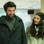 Engin Akyürek e Tuba Büyüküstün aparecem sorrindo e conversando em um ambiente interno durante uma cena da novela Kara Para Aşk.