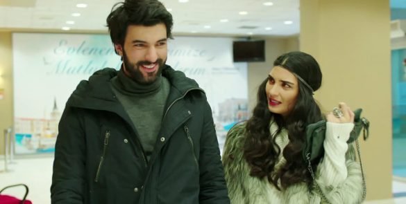 Engin Akyürek e Tuba Büyüküstün aparecem sorrindo e conversando em um ambiente interno durante uma cena da novela Kara Para Aşk.