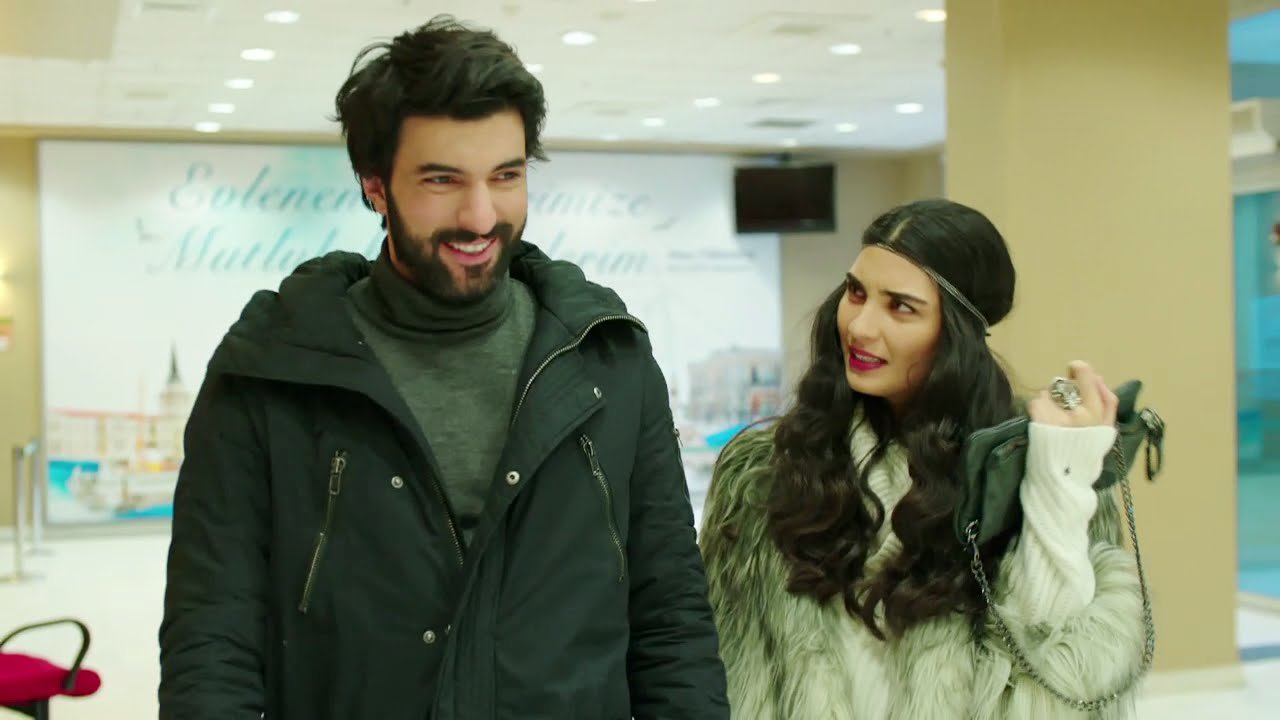 Engin Akyürek e Tuba Büyüküstün aparecem sorrindo e conversando em um ambiente interno durante uma cena da novela Kara Para Aşk.