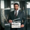 Can Yaman utilizando um terno em um escritório moderno, segurando uma claquete marcando o início da gravação da série Bro.