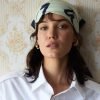 Pınar Deniz posa para um retrato usando uma camisa branca e um lenço estampado na cabeça, em frente a uma parede com papel de parede floral vintage.